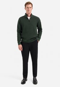 Man staat in een donkergroene gestructureerde sweater met kwart-rits, zwarte broek, zwarte laarzen, met één hand in zijn zak op een witte achtergrond.