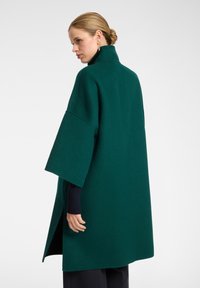 Abrigo de mezcla de lana verde con un alto cuello, mangas anchas y aberturas laterales. Textura suave, hasta la rodilla y diseño minimalista.
