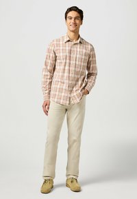 Camisa de manga larga a cuadros en beige con líneas rojas, cuello abotonado y bolsillo en el pecho. Combinada con pantalones de color claro y zapatos de ante.