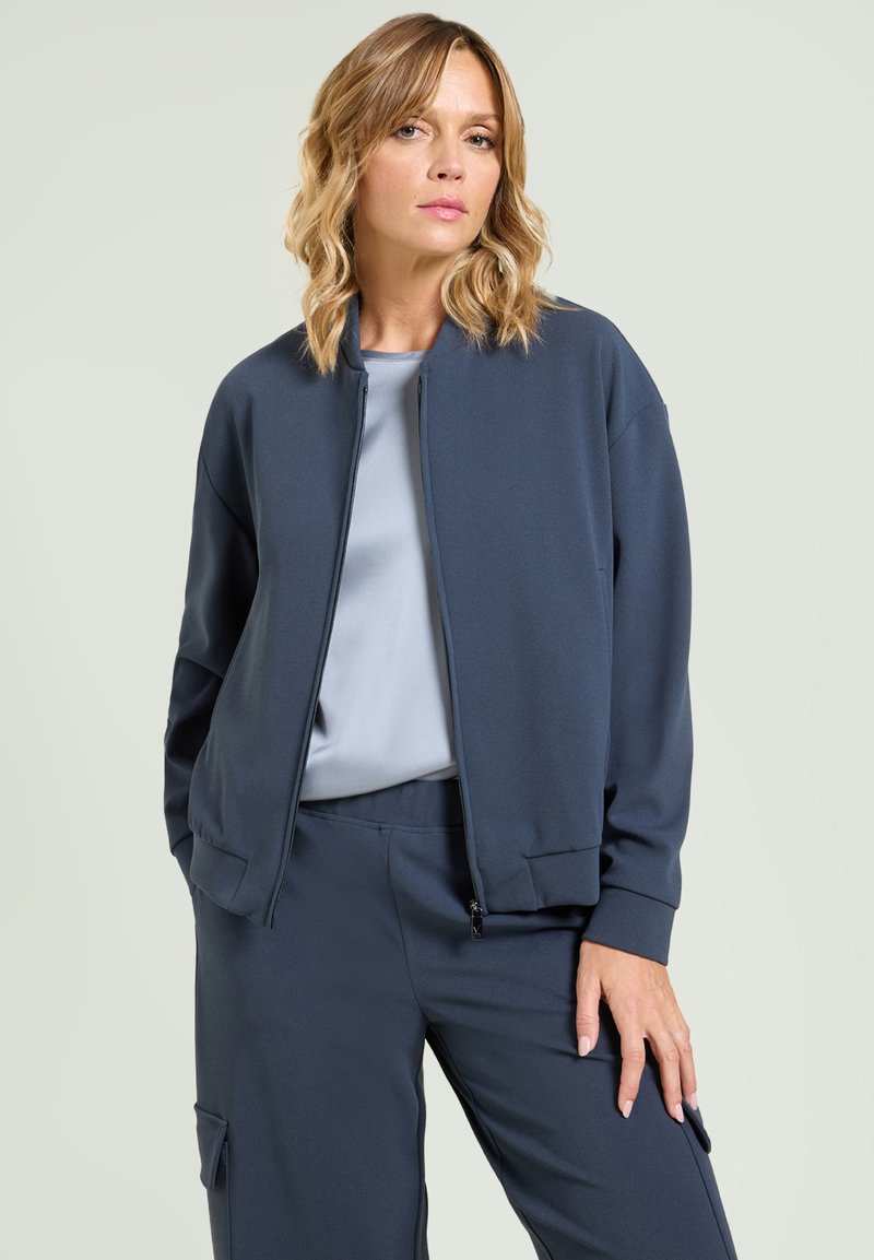 Donkerblauw bomberjack van zacht materiaal, met een ritssluiting, geribbelde manchetten en een ontspannen pasvorm, gecombineerd met een lichtblauw topje en bijpassende broek.