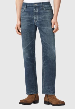 Person, der gerade geschnittene blaue Jeans und braune Wildlederstiefeletten trägt und vor einem neutralen Hintergrund steht.