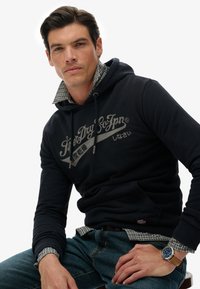 Granatowa bawełniana bluza z kapturem z logo z przodu, regulowanymi sznurkami i wystającą kołnierzykiem w kratę. Noszona z niebieskimi dżinsami i zegarkiem.