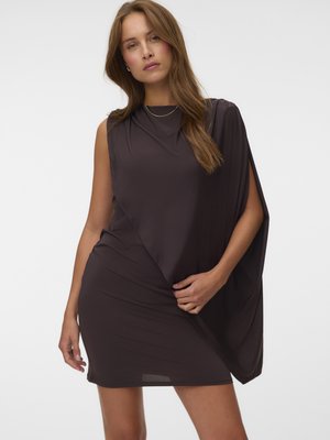 Femme portant une robe noire asymétrique sans manches avec un tissu drapé d'un côté et un petit collier en or sur un fond uni.