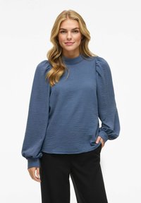 Kék, texturált, hosszú ujjú blúz magas nyakkivágással és puffos ujjakkal, fekete nadrággal párosítva. Puha anyag, laza fazon.