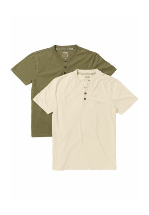 Lächelnder Mann trägt beige kurzärmliges Hemd, braune Hose und Schuhe, hält eine gefaltete karierte Jacke über der Schulter und steht vor einem schlichten Hintergrund.