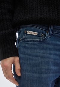 Σκούρο μαύρο πουλόβερ με ριμπ ύφανση συνδυασμένο με κλασικά μπλε τζιν της denim, με λογότυπο Calvin Klein στο μπροστινό τσεπάκι. Απαλή υφή.