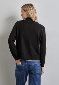 Maglione nero a maglia con colletto alto a coste, orlo e polsini a coste, indossato sopra jeans di denim azzurri chiari. Vista di spalle.