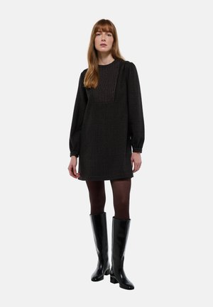 Robe noire texturée avec des manches bouffantes, dotée d'un panneau rayé et de boutons. Associée à des bottes noires jusqu'au genou et des collants sombres.