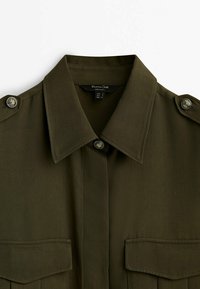 Olive green shirt with buttoned shoulder epaulettes, a folded collar, two chest pockets, and a smooth fabric texture.