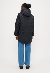 Črni prešiti dolg plašč s kapuco, z robom, ki se spušča navzdol, nošen prek svetlo modrih širokih hlač in črnih čevljev.