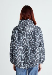 Moterys su pečių ilgio raudonais plaukais vilkinti pilką ir juodą leopardų rašto bliuzoną bei mėlynus džinsus, atsisukusi nuo objektyvo prieš paprastą šviesų foną.