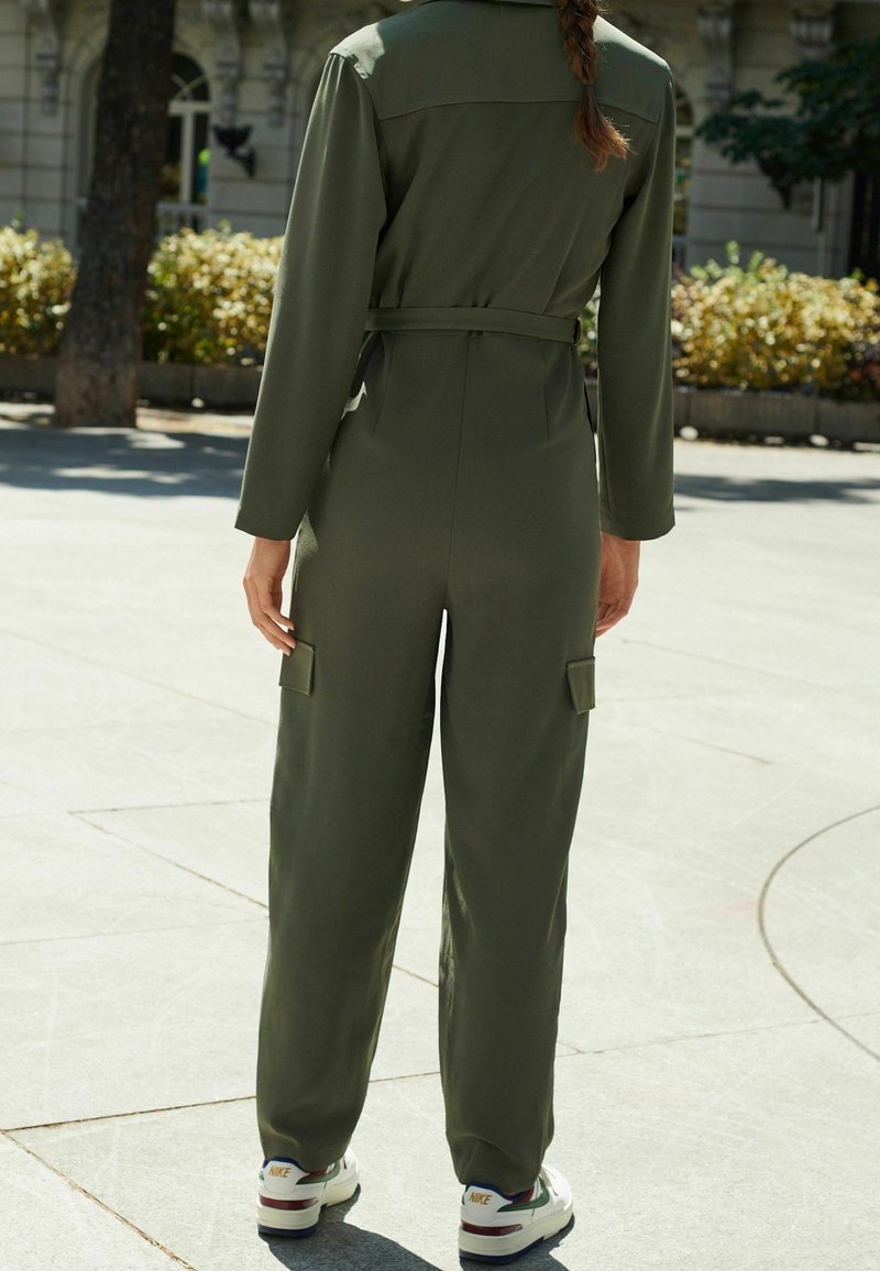 Personne avec une longue tresse portant une combinaison vert olive et des baskets blanches Nike, debout sur une surface pavée en plein air avec des arbres et des arbustes.