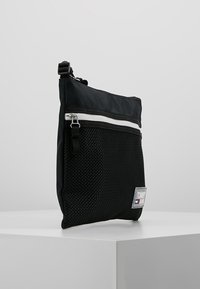 Borsa in nylon nera con fondo in rete, dotata di chiusura zip superiore e una striscia decorativa bianca. Include una toppa con logo e una tracolla regolabile.