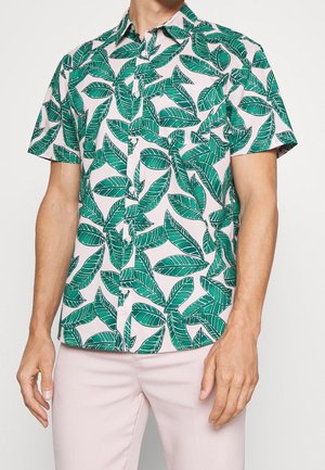 Chemise à manches courtes avec un motif de feuilles vertes sur un tissu rose clair, associée à un pantalon rose clair, portée par une personne debout.