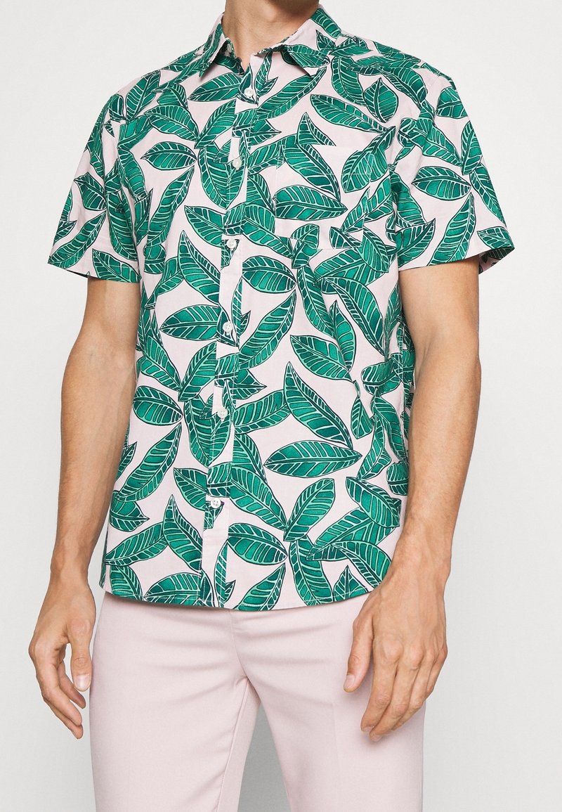 Camicia a maniche corte con un motivo di foglie verdi su un tessuto rosa chiaro, abbinata a pantaloni rosa chiaro, indossata da una persona in piedi.