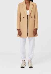 Cappotto in misto lana beige con un design sartoriale, bottoni doppi, e rever a punta, indossato su pantaloni felpati grigi e sneakers color camoscio.