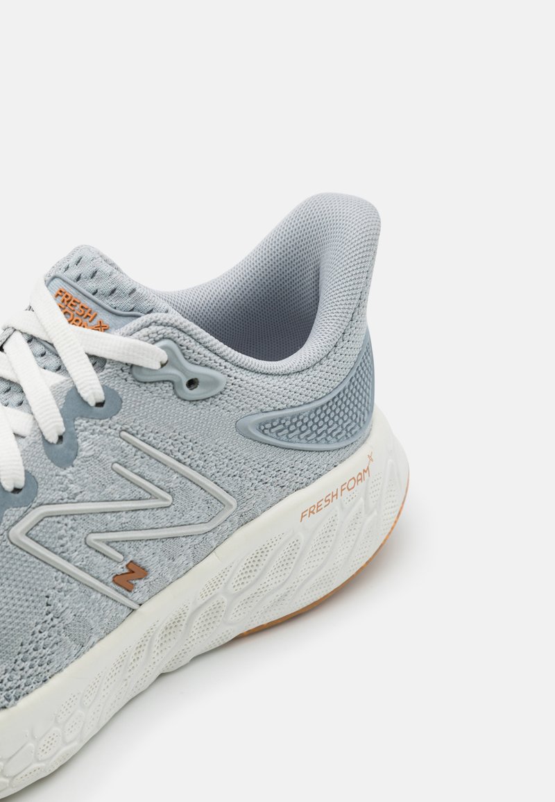 Lichtgrijze sportschoen met een bovenwerk van mesh, witte veters, een gepolsterde kraag, oranje accenten en een gestructureerde zool. Bevat "FRESH FOAM" branding.