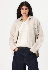 Beige košile s knoflíky, vyrobená z měkké látky, nošená přes krémový svetr s copánkovým vzorem, v kombinaci s tmavými džíny.