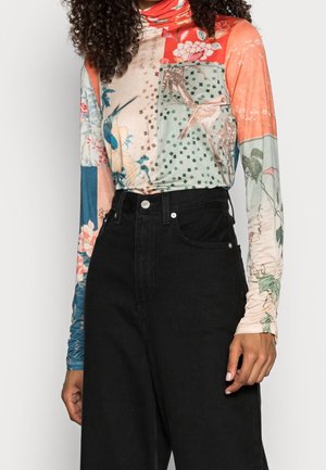 Femme portant un haut à col roulé à manches longues coloré avec des motifs d'oiseaux et de fleurs, associé à un pantalon noir taille haute.