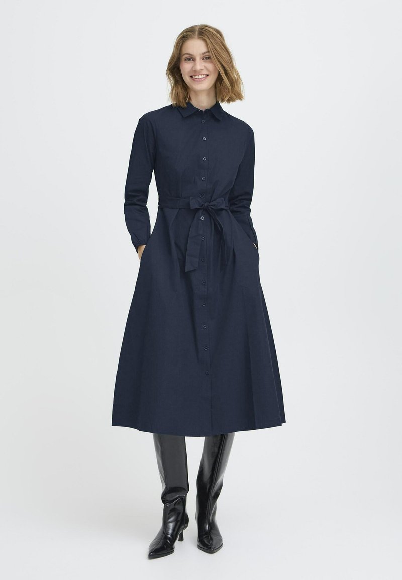 Navyblaues Hemdkleid aus Stoff mit Knopfverschluss vorne, langen Ärmeln, einem Bindegürtel in der Taille und seitlichen Taschen. Schwarze kniehohe Stiefel.