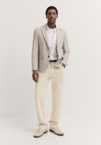 Hellgrauer Blazer über einem weißen Hemd, beigen Hosen und neutralen Sneakers. Auffällige Knopfdetails am Blazer; glatte Stoffstruktur.