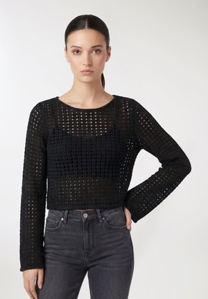 Giovane donna con lunghi capelli scuri raccolti, che indossa un top nero a maniche lunghe in crochet e jeans scuri a vita alta, in piedi con una mano sui fianchi.