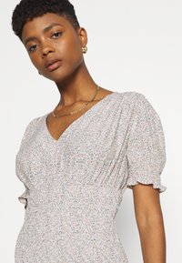 Vestido floral com decote em V, mangas curtas bufantes e cintura franzida. O tecido apresenta uma paleta de cores pastel suaves com pequenas flores multicoloridas.
