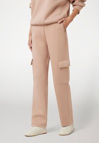 Šviesiai rožinės spalvos cargo džinsinės kelnės, pagamintos iš minkštos medžiagos, su šonine kišene ir apatiniais rankovėmis, derinamos su baltomis sportinėmis bateliais.