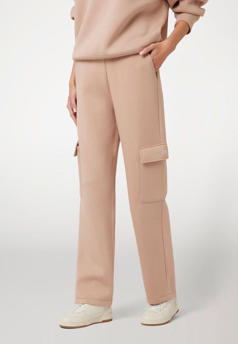 Šviesiai rožinės spalvos cargo džinsinės kelnės, pagamintos iš minkštos medžiagos, su šonine kišene ir apatiniais rankovėmis, derinamos su baltomis sportinėmis bateliais.