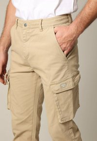 Beige cargobroek met een reguliere pasvorm, voorzien van zijzakken, een met een rits afsluitbare zak op het been en een merkembleem aan de zijkant. Gemaakt van katoen.