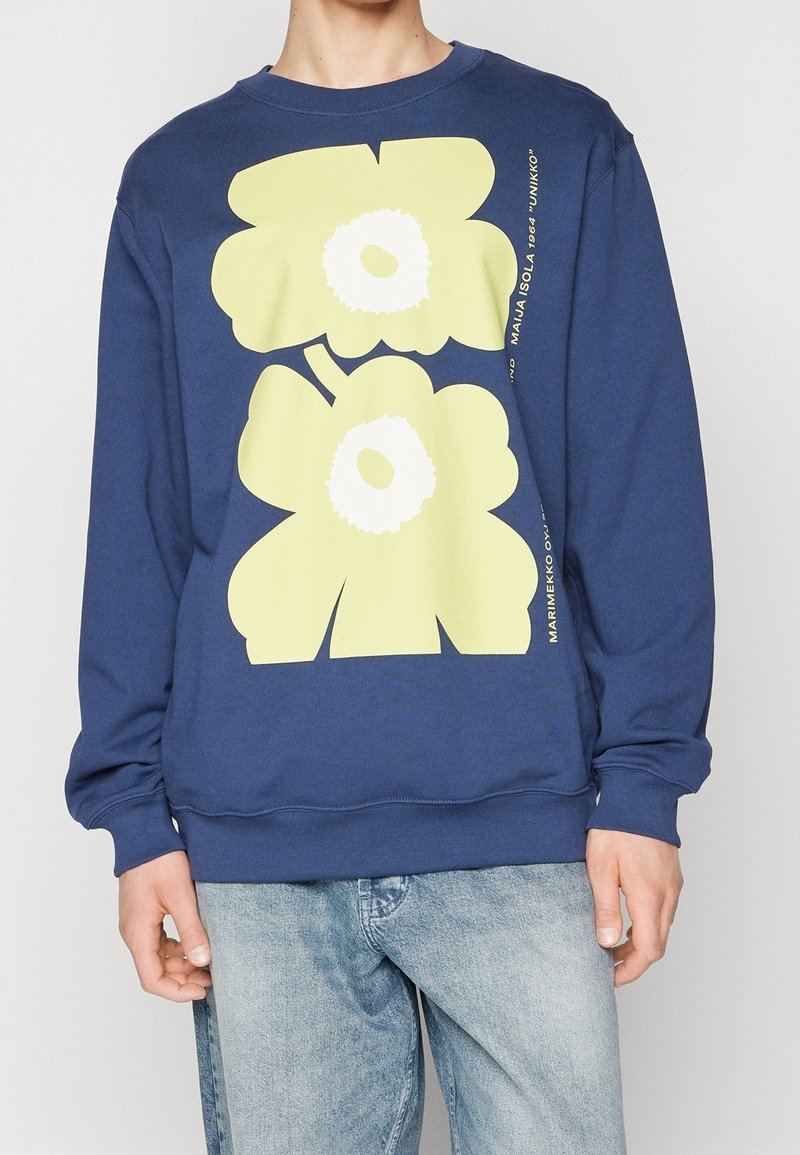 Námořnický modrý svetr s velkými žlutými květinovými potisky, kulatým výstřihem a žebrovanými manžetami a lemem. Tištěný text na boku. Pohodlný střih.
