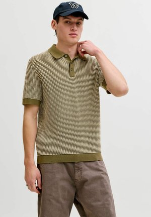 Mladý muž nosí vzorované béžovo-olivové polokošeľu, hnedé nohavice a čiernu šiltovku s bielym logom "W", stojí pred jednofarebným pozadím.