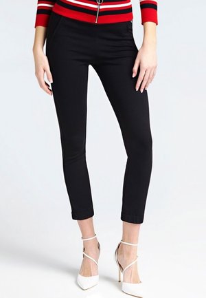 Frau trägt eng anliegende schwarze knöchellange Hose kombiniert mit weißen High Heels und einem rot-schwarz gestreiften Oberteil mit Reißverschluss.