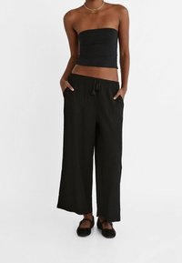 Zwarte strapless top en losse, zwarte broek van gekreukeld stof met een verstelbare taille met aantrekkoord. Uitgevoerd met twee zijzakken en platte schoenen.