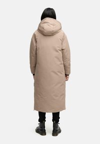 Lang beige pufferjakke med stor hætte, glat tekstur og sidelommer. Bæret over sorte bukser og støvler, set bagfra.