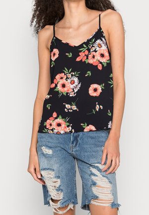 Femme portant un haut à bretelles spaghetti noir à fleurs roses et blanches, associé à un short en jean bleu déchiré.