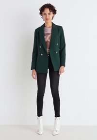 Zelený dvouřadý blejzr, černé skinny džíny, bílé kotníkové boty s akcenty. Blejzr má lesklé knoflíky a přizpůsobený design.