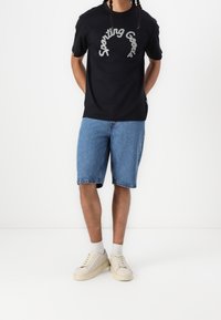 Mustat puuvillaiset t-paidat, joissa on valkoista tekstiä "Sporting Goods", yhdistettyinä sinisiin farkkushortseihin ja beigeihin lenkkareihin, joissa on teksturoitu pohja.