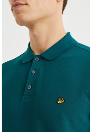 Mies pukeutunut petroolinvihreään poolopaitaan, jossa on kolme nappia ja pieni keltainen kirjailtu logo rinnassa, yksiväristä taustaa vasten.