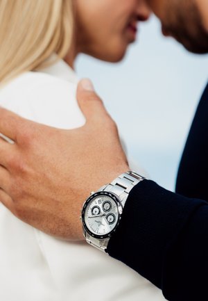 Montre chronographe argentée avec une lunette noire, trois sous-cadrans et un bracelet en maillons métalliques sur un poignet, partiellement obscurci par une main.