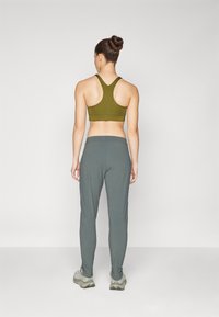 Olivegrüner Sport-BH mit Racerback-Design, kombiniert mit grauen Sporthosen im lässigen Schnitt und mit Bündchen an den Säumen. Das Modell trägt graue Sneakers.