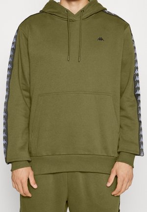 Mann trägt einen olivgrünen Kapuzenpullover mit schwarz-weißen, gemusterten Streifen an den Ärmeln und einer Kängurutasche vorne.