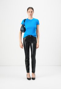 Vrouw in een felblauw, kort-gevleugeld topje, zwarte leren broek, zwarte hakken en een kleine zwarte schoudertas, staand tegen een witte achtergrond.
