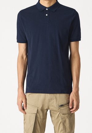 Mann trägt ein schlichtes dunkelblaues kurzärmeliges Polohemd und beige Cargohosen mit Taschen und Reißverschlüssen, steht vor weißem Hintergrund.