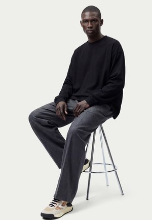 Man draagt een zwart langarmig shirt, grijze broek en witte sneakers, zit op een metalen kruk tegen een effen achtergrond.