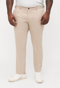 Beige tailored trousers with a smooth texture, straight fit, front pockets, and a single button closure, paired with white sneakers.