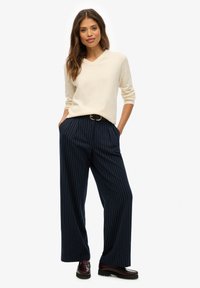 Crèmekleurige gebreide trui met een V-hals, gecombineerd met marineblauwe pantalon met streepjes en donkerburgundy loafers. De pantalon heeft een riem.