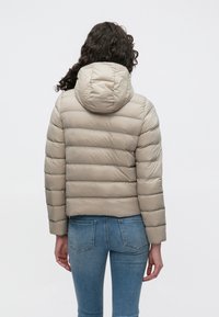 Béžová prešívaná bunda s kapucňou, s horizontálnou prešívanou textúrou a skrátenou dĺžkou, kombinovaná s modrými džínsami z denimu.