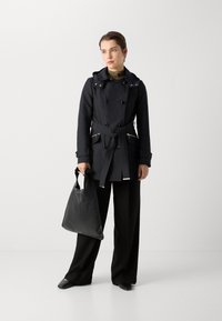 Marineblauwe trenchcoat met capuchon, dubbelzijdig ontwerp, riem en met rits afgesloten zakken, gecombineerd met zwarte wijde broek en een zwarte totebag.