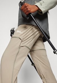 Beige välispüksid tekstureeritud viimistlusega, varustatud lukuga taskute ja Jack Wolfskin logo. Koos musta matkakepiga.